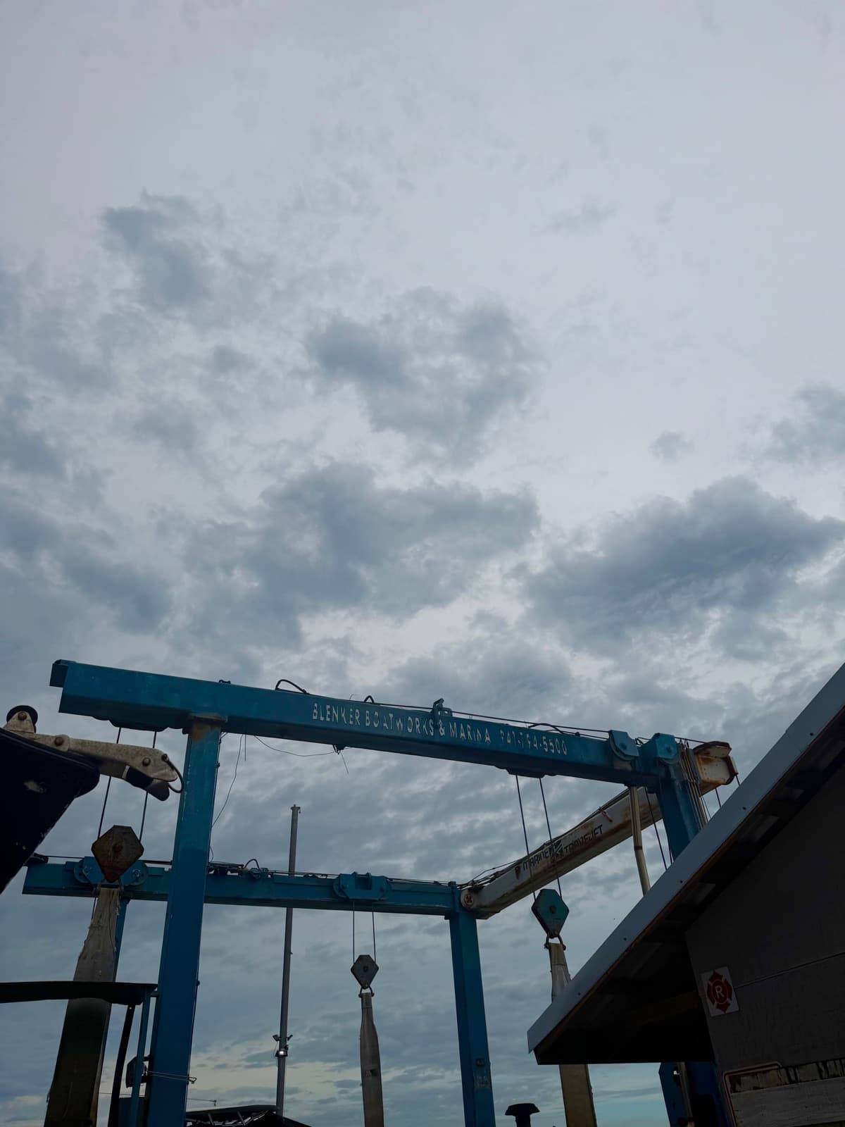 The blue steel frame of the yard's travel lift against a cloudy sky, with the business name painted along the top beam and lifting slings hanging below