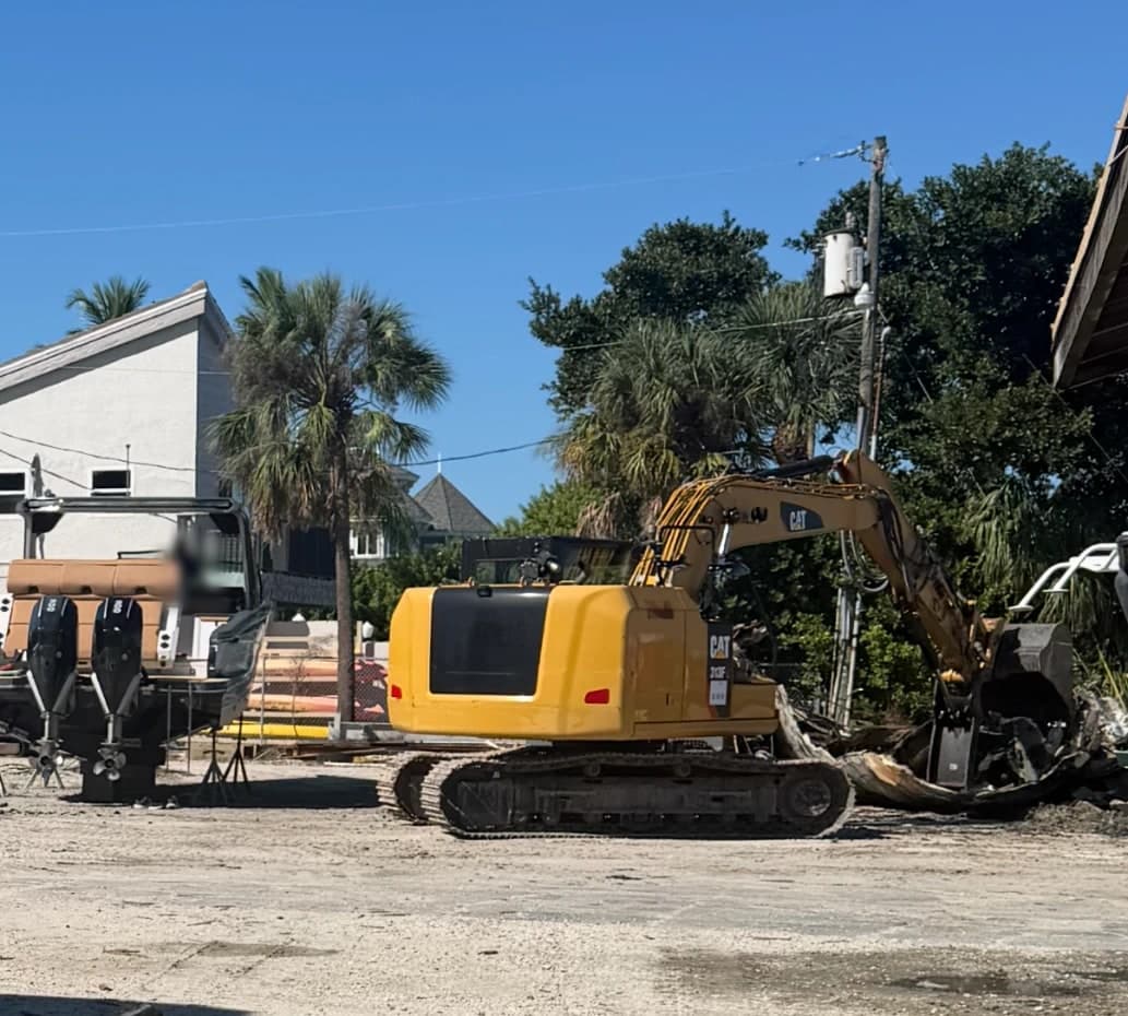 An excavator breaking up an old boat hull in a gravel boat yard, beside a boat with two outboard motors on stands, with palm trees behind