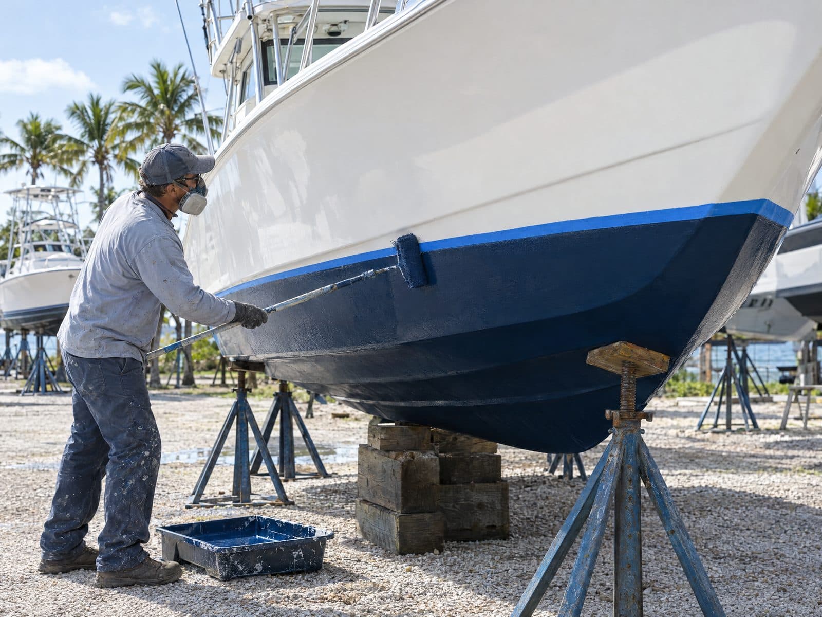 A yard worker in a respirator rolling fresh dark blue antifouling paint onto the hull of a fishing boat on jack stands, a taped waterline above the new paint