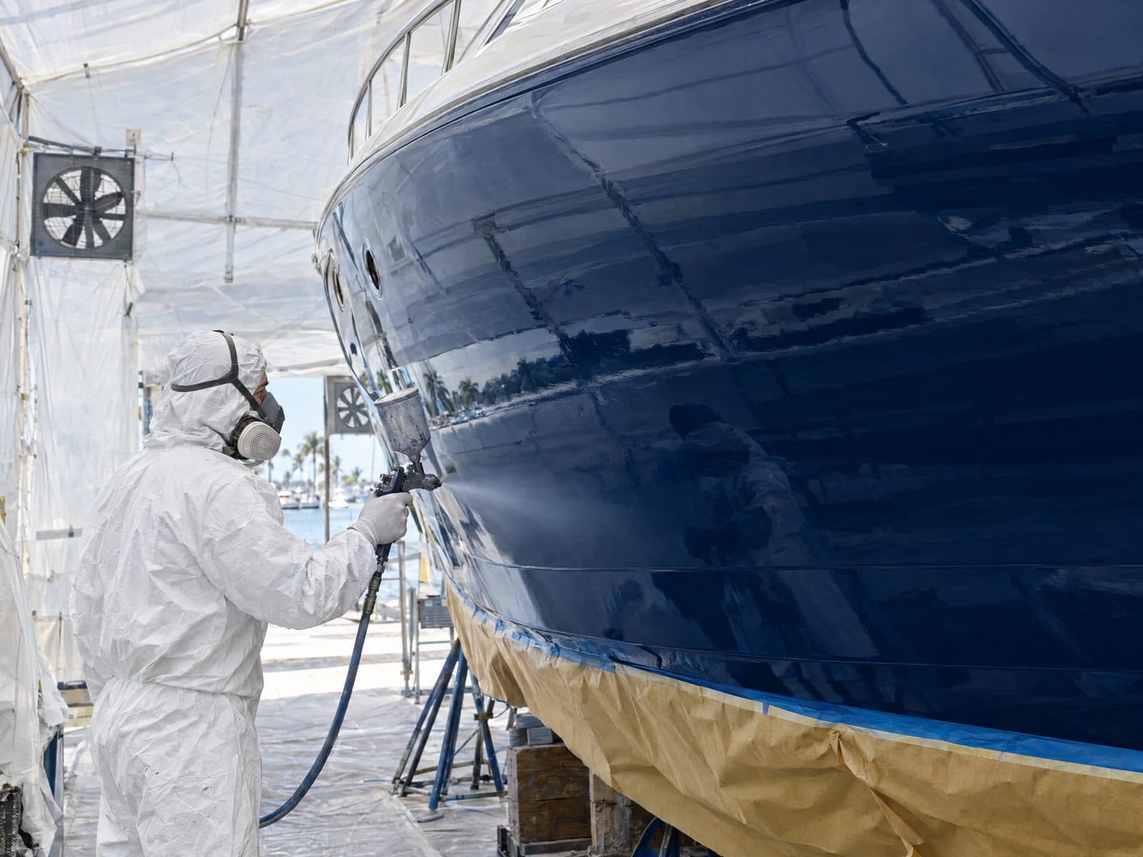 A painter in a white protective suit spraying a glossy navy blue finish onto a boat hull inside a ventilated paint tent